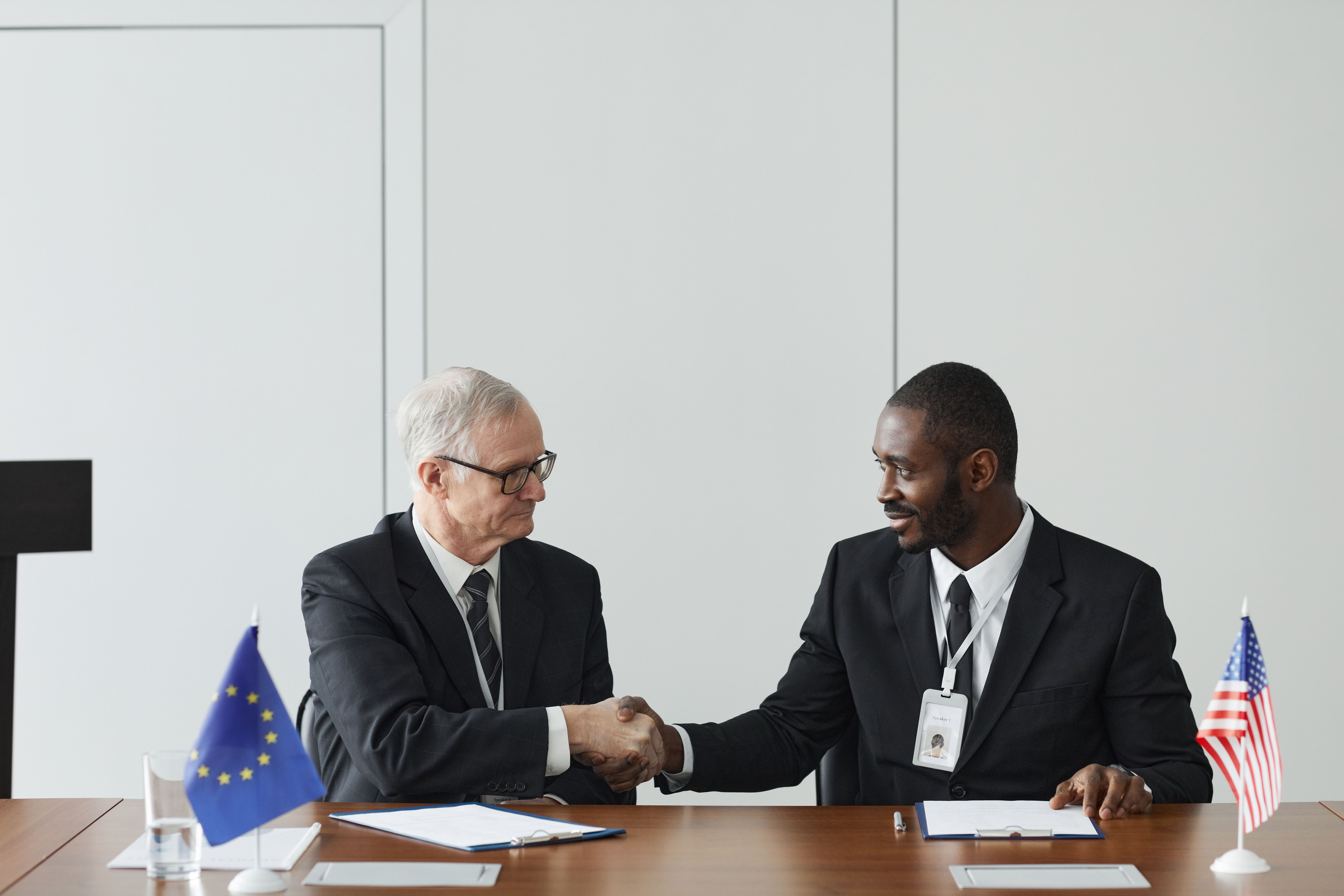 Corporate handshake between diverse businessmen with EU and US flags symbolizing cross-border legal partnership